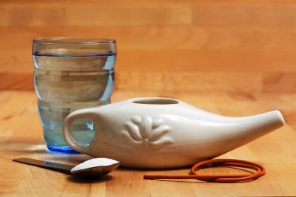 A neti pot sitting next to a glass and some nasal rinse solution on a teaspoon
